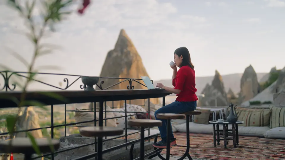Woman sat working on a laptop whilst sat on a rooftop terrace overlooking a mountain