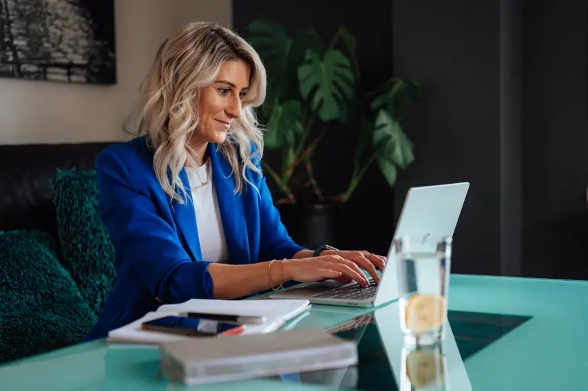 Blonde woman in a blue blazer working on laptop at kitchen table